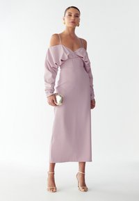 Fialové šaty s rukávy s odhalenými rameny, vypasovaným pasem a rovnou sukní. Modelka drží metalickou kabelku a má na sobě sandály na podpatku.