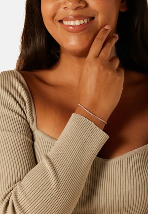 Zilveren armband met een delicaat ontwerp, voorzien van kleine edelstenen. Draagt aan een gebruinde pols, hand rustend bij de kin, tegen een neutrale achtergrond.