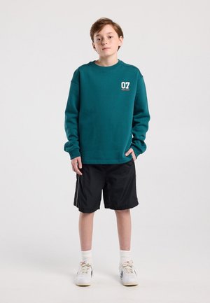Jonge jongen staand, gekleed in een turkooise sweatshirt met logo "07", zwarte korte broek, witte sportsokken en witte sneakers tegen een effen lichte achtergrond.