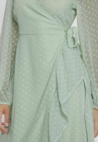 Lys grønn omslagskjole laget av teksturert stoff med hvite prikker. Har lange gjennomsiktige ermer og en knyttet midjedetalj som skaper en lagdelt effekt.