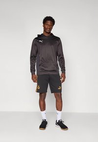 Musta urheilullinen huppari hopeanvärisellä logolla, yhdistettynä mustiin shortseihin, joissa on oransseja yksityiskohtia. Mallilla on valkoiset sukat ja mustat tennarit.