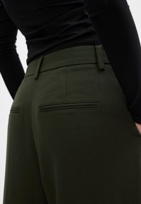 Κοντινή λήψη ενός ατόμου που φοράει σκούρους ελαιόχρυνους παντελόνες με πίσω τσέπες και μαύρο εφαρμοστό μακρυμάνικο μπλουζάκι, εστιασμένο από τη μέση και κάτω.