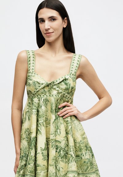 Γυναίκα με μακριά σκούρα μαλλιά φοράει αμάνικο, ανοιχτό πράσινο φόρεμα με λεπτομερή βοτανικό και αρχιτεκτονικό σχέδιο, ποζάροντας με το ένα χέρι στη μέση.
