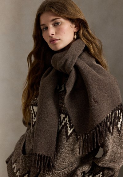 Brun ullskjerf med frynser, drapert rundt modellens hals, kombinert med en brun mønstret genser med kremfargede detaljer.