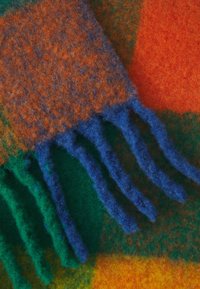 Gros plan d'une écharpe en laine multicolore avec des franges bleues et vertes sur un fond en tissu à motifs orange, vert et jaune.