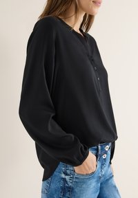 Blouse noire à manches longues avec un col à boutons et des poignets élastiques, associée à un jean en denim bleu clair déchiré. Texture lisse.
