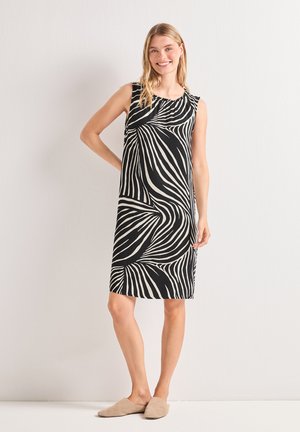 Femme souriante portant une robe noire sans manches longueur genou avec un motif abstrait en spirale blanche et des chaussures beiges à enfiler, debout contre un mur clair.