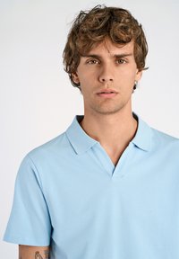 Ljust blå polo med kort krage, tillverkad av ett mjukt och slätt tyg. Har en klassisk passform och en subtil textur.