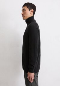Schwarzer Reißverschluss-Sweater mit hohem Kragen, gerippten Bündchen und Saum. Weiche Textur und taillierte Passform, die von der Seite sichtbar ist.