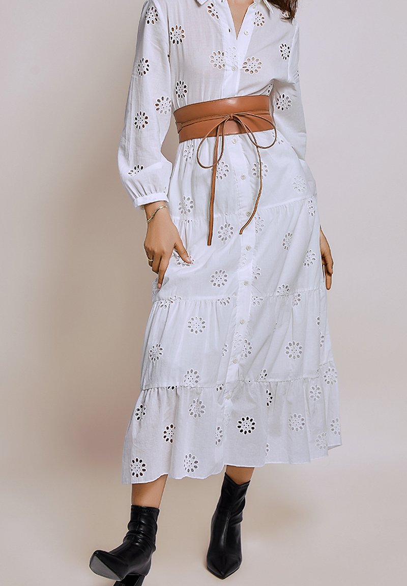 Witte katoenen jurk met bloemen-oeillet-details, knopen aan de voorkant, gelaagde zoom en gegolfde mouwen, gecombineerd met een bruine leren riem en zwarte enkellaarsjes.