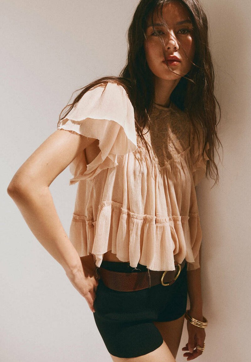 Femme aux longs cheveux foncés portant une blouse beige ample et un short noir avec une ceinture marron, debout devant un fond clair uni.