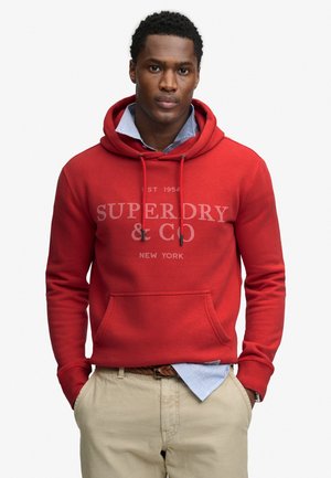 Ung mand iført en rød Superdry-hoodie over en blå skjorte med krave og beige bukser, stående med hænderne i lommerne mod en hvid baggrund.