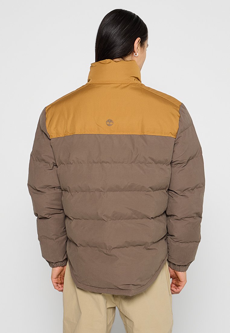 Giacca imbottita marrone con sezione superiore a costine color sabbia. Presenta sezioni cucite e un logo sul retro. Design leggero e trapuntato.