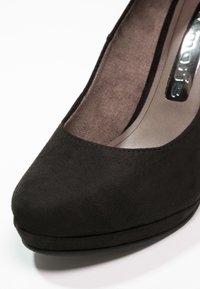 Chaussure à talon haut en suède noir avec un bout arrondi et une légère plateforme. L'intérieur présente un revêtement en brun doux et un branding en argent minimaliste.