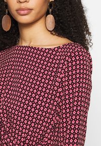 Robe à motifs rouges et noirs avec des formes de diamant, présentant un décolleté large et un tissu drapé. De grandes boucles d'oreilles en bois en forme de cercle complètent le look.