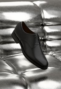 Chaussure de ville en cuir noir avec une texture lisse, bout rond et design à lacets. Mise en scène sur un fond argenté brillant.