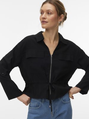 Frau trägt eine schwarze, langärmlige Jacke mit Reißverschluss, Kordelzug in der Taille und zwei Brusttaschen, kombiniert mit blauen Jeans, Hände in die Hüften gestützt.