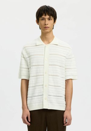 Fiatal férfi, aki fehér rövid ujjú, csíkos, nyitott kötésű mintázatú ingben és sötét nadrágban áll egyszínű háttér előtt.