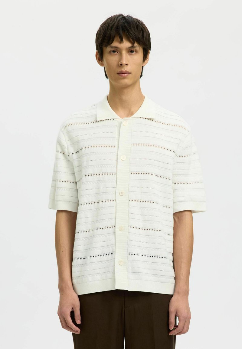 Jeune homme portant une chemise blanche à manches courtes avec un motif rayé en maille ajourée et un pantalon sombre, debout devant un fond uni.