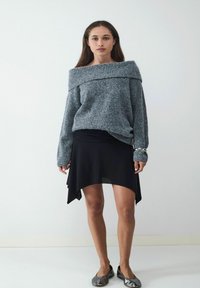 Grijze off-shoulder trui met geribbelde halslijn, gecombineerd met een zwarte asymmetrische rok. Model draagt grijze platte schoenen. Minimalistische achtergrond.