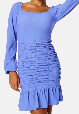 Woman wearing a fitted light blue dress with long sleeves, square neckline, ruched midsection, and ruffled hem standing against white background.