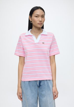 Vrouw draagt een roze-wit gestreept poloshirt met een witte kraag en blauwe spijkerbroek, staand tegen een effen witte achtergrond.