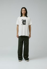 Vit t-shirt med en svart grafisk tryck som läser "FORMER" på bröstet, i kombination med mörkgröna vida byxor och svarta skor.