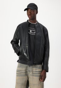 Jachetă din piele neagră cu guler înalt, detalii cu cusături albe și fermoar frontal; combinată cu un tricou grafic negru și pantaloni din denim deschis.
