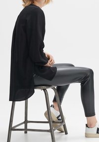 Femme assise sur un tabouret en métal porte un chemisier noir à manches longues, un pantalon en cuir noir et des baskets blanches avec des accents noirs.