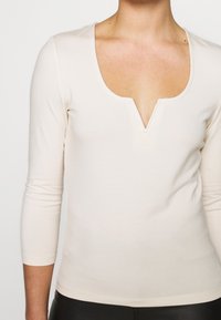 Beige långärmad topp med djup V-hals och lätt stretch, gjord av mjukt tyg. Har en figursydd silhuett och slät textur.