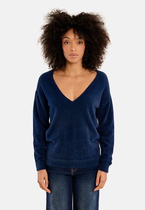 Donna con capelli ricci che indossa un maglione a V blu navy peloso e jeans blu scuro, in piedi davanti a uno sfondo bianco uniforme.