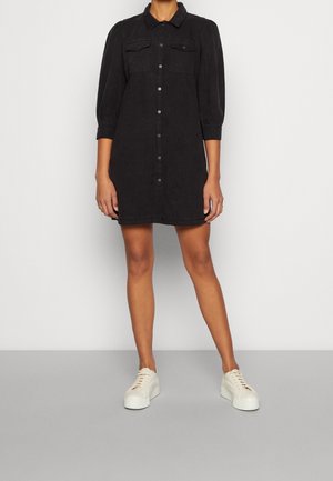 Kvinde iført en sort skjortekjole med knapper, puffede ærmer og hvide plateau-sneakers, stående mod en ensfarvet lys baggrund.