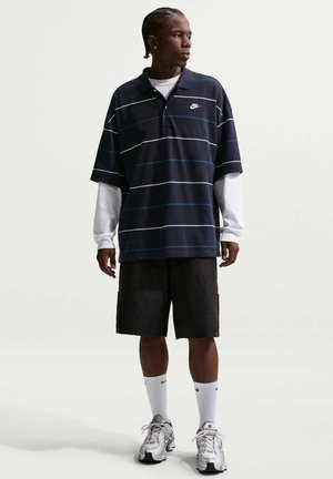 Jeune homme aux cheveux tressés portant un polo Nike à rayures foncées sur une chemise blanche à manches longues, un short noir, des chaussettes blanches Nike et des baskets argentées.