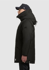 Černá zateplená parka s vysokým límcem, kapucí na stažení, zipovou kapsou, nášivkou s logem na rukávu a mírně zakřiveným lemem.