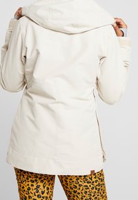 Helle beige wasserfeste Jacke mit Kapuze, die eine strukturierte Oberfläche und Nahtdetails aufweist, kombiniert mit gemusterten gelben und schwarzen Hosen.