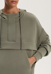 Veľká olivovo zelená mikina s textúrovaným povrchom, s vysokým golierom, šnúrkami a veľkým predným vreckom.