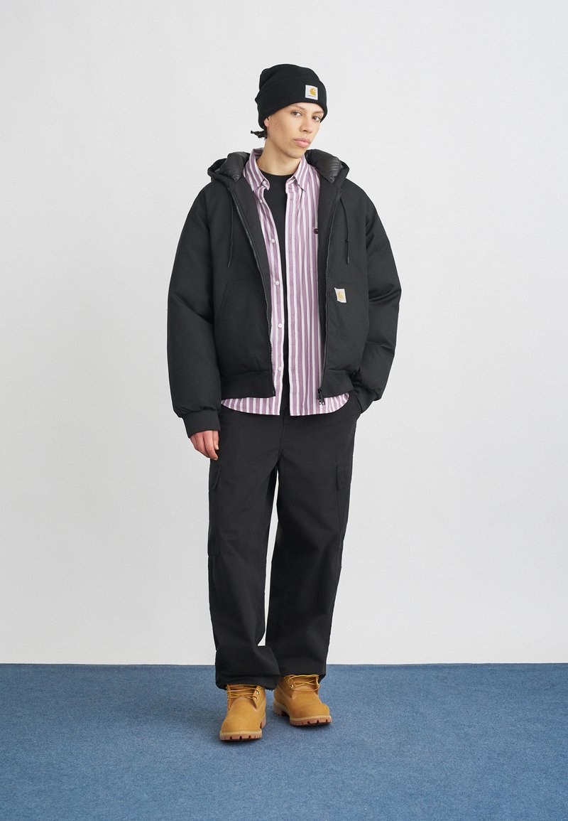 Chaqueta acolchada negra con cremallera y forro gris, superpuesta a una camisa de botones a rayas. Pantalones oscuros y botas de trabajo marrones completan el atuendo.