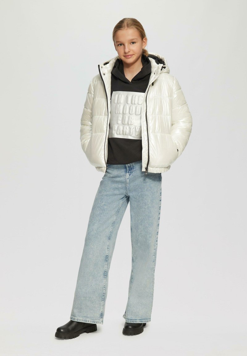 Witte, glanzende, gewatteerde jas met capuchon, gedragen over een zwarte hoodie met reliëftekst. Gekozen in combinatie met lichtblauwe wijde jeans en zwarte laarzen.