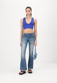 Blå ribbet crop topp, flagrende lysvaskede jeans, svarte plattform sandaler, og en liten blå håndveske. Modell står mot en hvit bakgrunn.