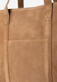 Bruine suede schoudertas met twee stevige handvatten, voorzien van een voorvak en nette stiksels aan de zijkanten voor extra detail.