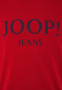 Tissu rouge avec un texte noir en gras indiquant JOOP! JEANS centré sur le matériau.