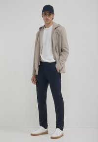 Giacca felpata beige, t-shirt bianca, pantaloni blu navy e sneakers bianche con suola color sabbia. Modello in piedi su uno sfondo neutro.