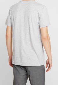 Osoba v krátkém rukávu svetru světle šedé barvy s melírem a šedých kalhotách, zobrazená zezadu na jednobarevném pozadí.