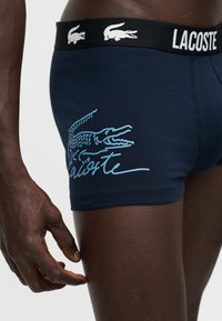 Άτομο που φοράει μπλε σκούρο μποξεράκι Lacoste με μεγάλο ανοιχτό γαλάζιο λογότυπο κροκόδειλου και επιγραφή στο πλάι, μαύρη ζώνη μέσης με branding.