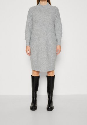 Kobieta w szarej, dzianinowej sukience swetrowej do kolan, zestawionej z czarnymi, skórzanymi kozakami do kolan, stojąca na tle jednolitego białego tła.