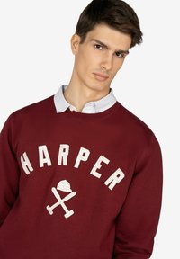 Sudadera burdeos con "HARPER" en texto blanco y un logotipo gráfico. Presenta un cuello redondo y un look en capas con un collar de rayas debajo.