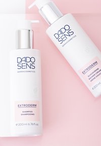 Two white cosmetic bottles with pump tops on a pink background. Labels read "DADO SENS EXTRODERM" for shampoo and shower cream.