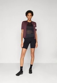 Purpurová cyklistická bunda s krátkymi rukávmi cez čierne priliehavé tričko, v kombinácii s čiernymi šortkami a cyklistickými topánkami; bunda má biely akcentný pás.