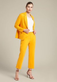 Geel blazer en bijpassende broek tot de enkel van een gladde stof. De blazer heeft een geknipt revers; gedragen over een witte top en gecombineerd met bruine hakken.