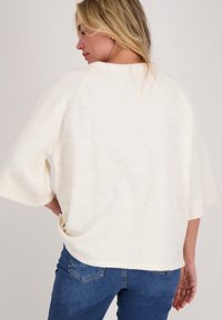 Licht beige trui met een losse pasvorm, korte mouwen en een ronde halslijn. Zachte textuur met een casual ontwerp, gecombineerd met blauwe jeans.